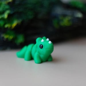 Puede incluir: Una pequeña figura de dinosaurio verde con púas blancas y ojos negros. El juguete está tumbado sobre una superficie clara, con un fondo borroso de follaje verde y madera oscura.