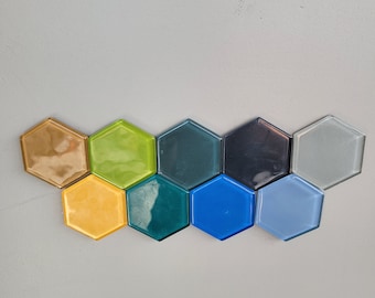 Op de afbeelding: Een set van negen zeshoekige tegels in verschillende kleuren, waaronder goud, groen, teal, blauw en grijs. De tegels zijn in een halve cirkelvorm gerangschikt.
