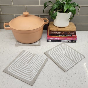 Puede incluir: Dos posavasos de cerámica con estampado blanco y gris, con fondo blanco y diseño geométrico. Los posavasos están sobre una encimera blanca con una maceta de color melocotón y una planta verde en el fondo.