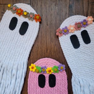 Könnte beinhalten: Drei weiße und rosafarbene, geisterförmige Wandbehänge mit gehäkelten Details. Jeder Geist trägt eine Blumenkrone aus bunten Garnblumen. Die Geister hängen an einer Holzwand.