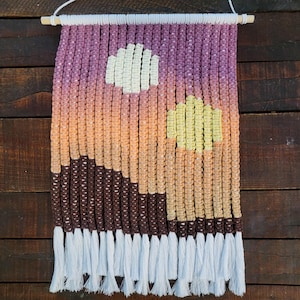 May include: A wall hanging made of yarn with a sunset scene. The scene features a brown mountain range, a yellow sun, and a white sun. The hanging has white tassels at the bottom.