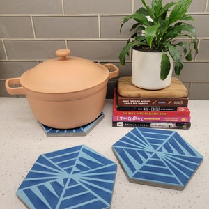 May include: Four blue and grey hexagonal trivet coasters with a geometric design. The coasters are on a white worktop with a pink pot and a houseplant in the background.