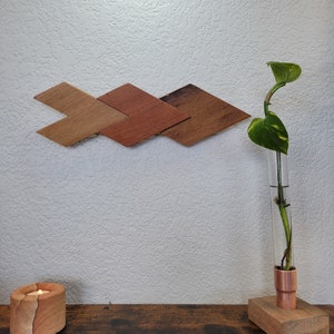 May include: A wooden wall decor piece in the shape of three connected diamonds. The diamonds are painted in shades of brown, orange, and red. A small potted plant with green leaves is in a copper vase on a wooden stand. A candle is lit in a stone holder on a wooden table.
