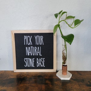 Puede incluir: Un letrero de madera con el texto "Pick your natural stone base" sobre un fondo de fieltro negro. Una planta verde en maceta está en un tubo de vidrio con una base de cobre y un soporte hexagonal blanco.