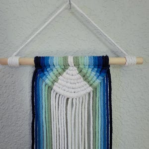 May include: A white, blue, and green macrame wall hanging with a wooden dowel at the top. The hanging is in the shape of a half-circle with fringe on the bottom.