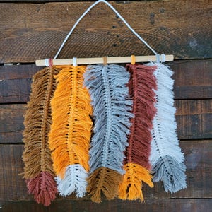 Op de afbeelding: Een macramé wandhanger met zes veervormige ontwerpen in tinten bruin, oranje, grijs en wit. De veren zijn bevestigd aan een natuurlijke houten deuvel en opgehangen met een wit koord. De achtergrond is een donker houten oppervlak.