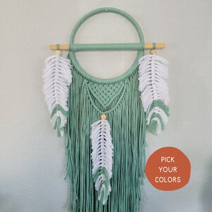 Puede incluir: Un atrapasueños de macramé verde y blanco con plumas colgando. El atrapasueños está hecho con un aro de madera y una varilla de madera. El texto "PICK YOUR COLORS" es visible en la esquina inferior derecha de la imagen.