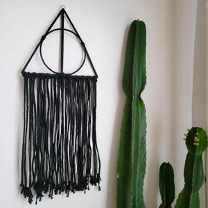 May include: Black macrame wall hanging with a geometric triangle shape and a circle in the center. The macrame is made with black cord and hangs from a gold hook.