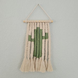 Pode incluir: Uma tapeçaria de macramé de parede com um design de cacto verde e uma barra de madeira na parte superior. A tapeçaria tem uma franja de borlas na parte inferior.