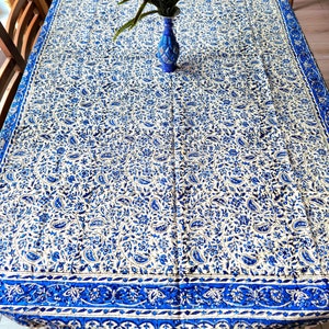 Può includere: Una tovaglia bianca con un motivo paisley blu. La tovaglia ha un bordo blu e frange. Un vaso con fiori viola è sul tavolo.