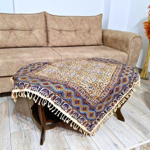 May include: A decorative, patterned table cover with a cream-colored background and a blue, yellow, and red border. The cover is fringed and rests on a dark wooden table. The table is in front of a beige sofa.