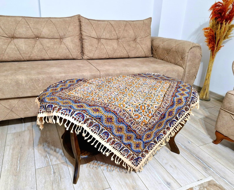 May include: A decorative, patterned table cover with a cream-colored background and a blue, yellow, and red border. The cover is fringed and rests on a dark wooden table. The table is in front of a beige sofa.