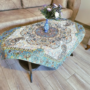 May include: A round, white table cloth with a blue and gold floral pattern. The table cloth is draped over a wooden table with a blue and white vase with flowers on top.