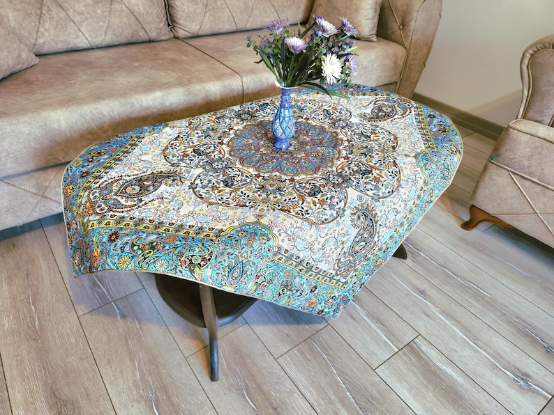 May include: A round, white table cloth with a blue and gold floral pattern. The table cloth is draped over a wooden table with a blue and white vase with flowers on top.