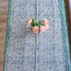 Può includere: Una tovaglia rettangolare con sfondo bianco e motivo paisley blu e verde. Il bordo è verde con un disegno floreale. Un mazzo di rose rosa è appoggiato sulla tovaglia.