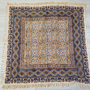 May include: A square, cream-colored textile with an intricate pattern. The central design features floral and bird motifs in shades of orange, blue, and pink. The border is a detailed geometric pattern in blue and gold, with a fringed edge.