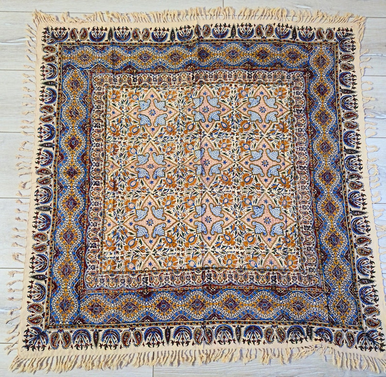 May include: A square, cream-colored textile with an intricate pattern. The central design features floral and bird motifs in shades of orange, blue, and pink. The border is a detailed geometric pattern in blue and gold, with a fringed edge.