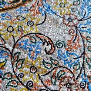 May include: Close-up of a silver fabric with a paisley pattern. The pattern is embroidered with colorful threads in shades of blue, green, orange, and yellow.