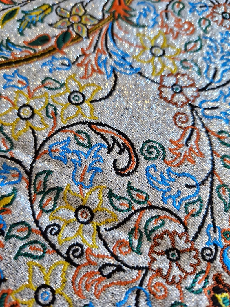 May include: Close-up of a silver fabric with a paisley pattern. The pattern is embroidered with colorful threads in shades of blue, green, orange, and yellow.