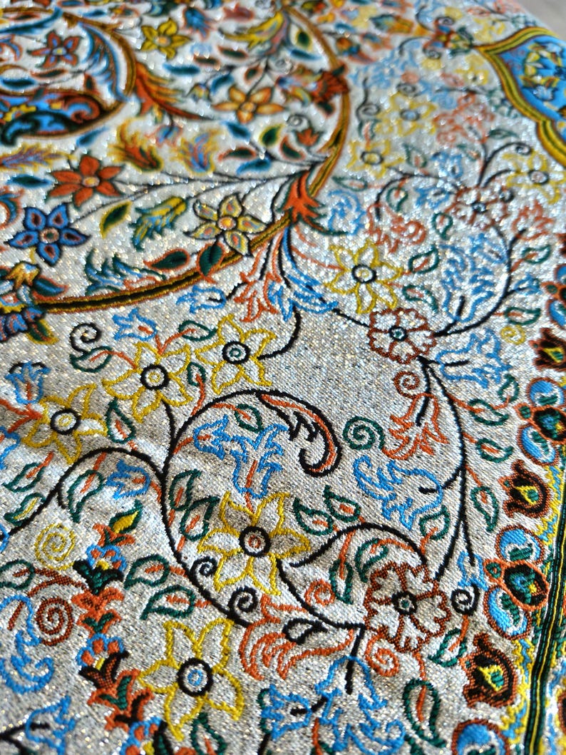 May include: Close-up of a white fabric with a colorful floral pattern. The fabric has a shiny, glittery texture. The pattern includes blue, green, orange, and yellow flowers.