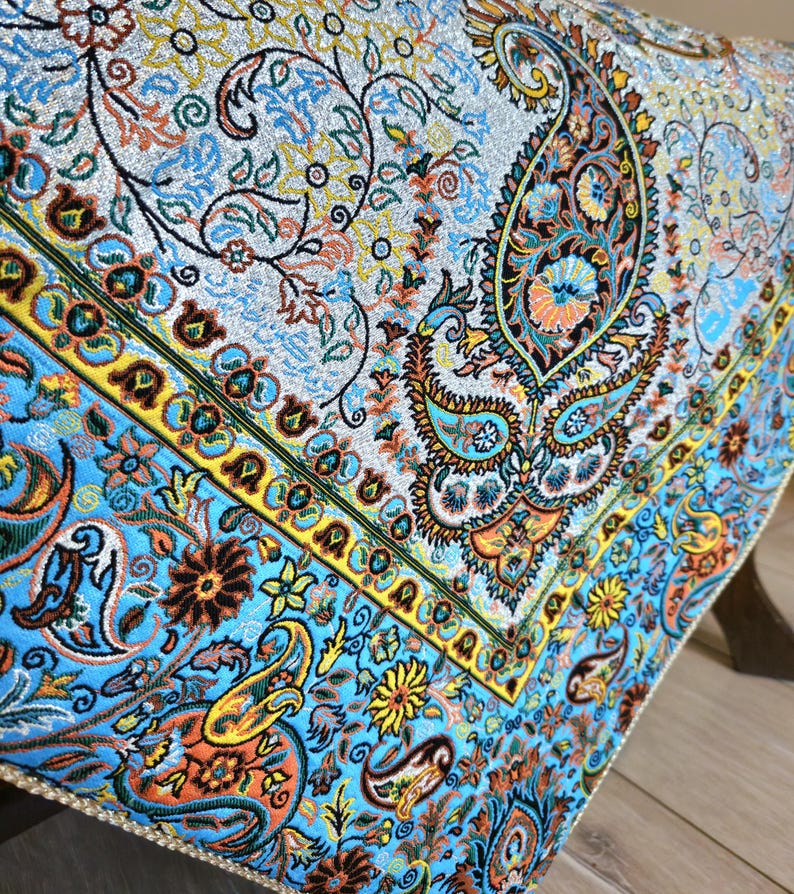 May include: A blue and gold patterned tablecloth with a paisley design. The tablecloth has a fringed edge and is made of a soft, luxurious fabric.
