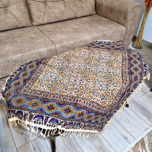 May include: An oval-shaped, patterned table cover with a cream-colored fringe. The cover features a floral design in shades of blue, yellow, and orange on a cream background. The table is dark brown and has four legs.