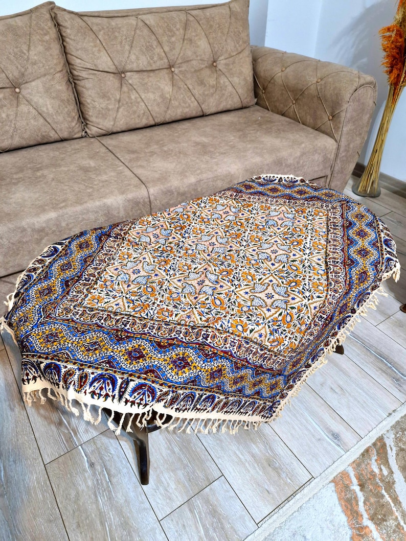 May include: An oval-shaped, patterned table cover with a cream-colored fringe. The cover features a floral design in shades of blue, yellow, and orange on a cream background. The table is dark brown and has four legs.
