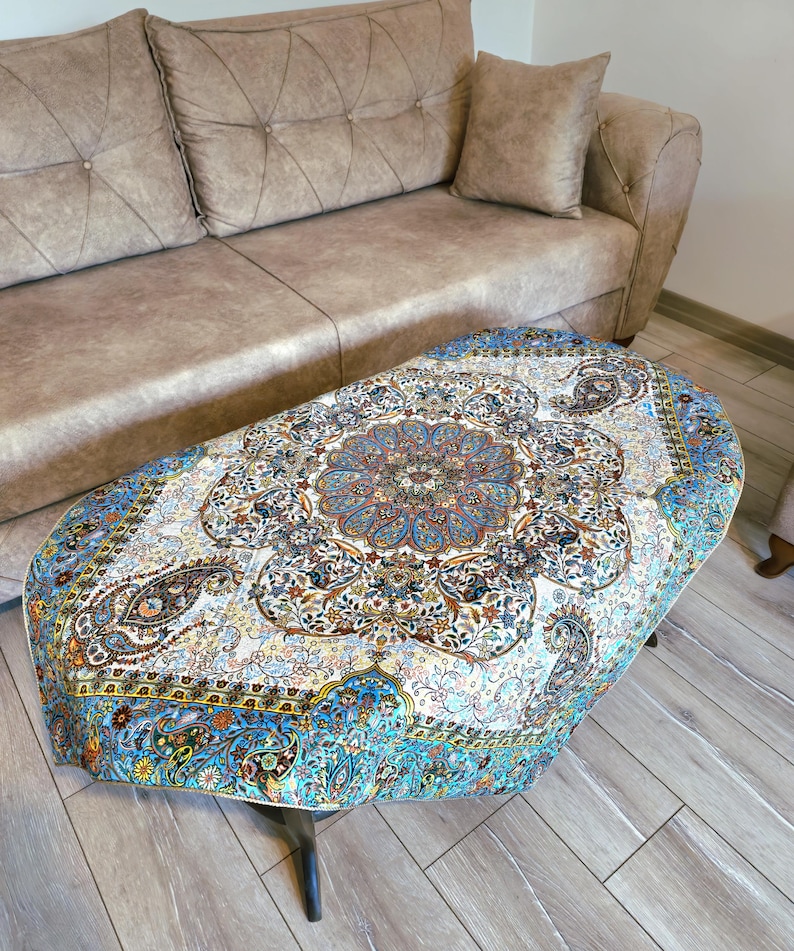 May include: A round, blue and white table cover with a floral and paisley design. The table cover is on a coffee table in front of a brown couch.