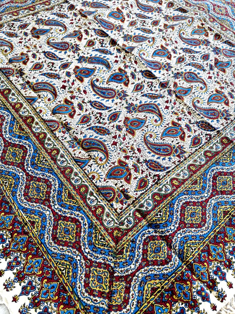 May include: A white tablecloth with a paisley pattern in shades of blue, red, and yellow. The tablecloth has a border with a repeating pattern of blue, red, and yellow.