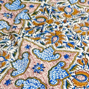 May include: Close-up of a fabric with a repeating pattern of stylized flowers, birds, and leaves in blue, gold, and green on a cream background. The design includes pink and gold accents.