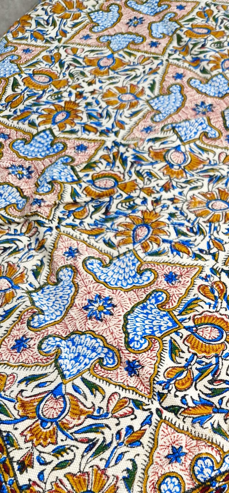 May include: Close-up of a fabric with a repeating pattern of stylized flowers, birds, and leaves in blue, gold, and green on a cream background. The design includes pink and gold accents.