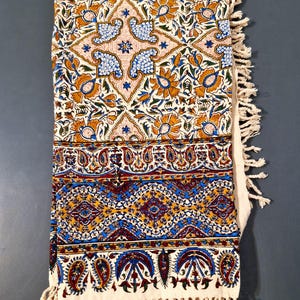 May include: A folded, cream-colored textile with intricate floral and paisley patterns in shades of orange, blue, and burgundy. The textile has a fringed edge and is likely a decorative throw or tablecloth.