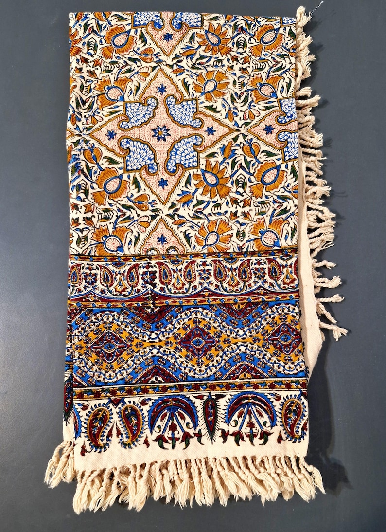 May include: A folded, cream-colored textile with intricate floral and paisley patterns in shades of orange, blue, and burgundy. The textile has a fringed edge and is likely a decorative throw or tablecloth.