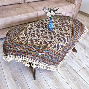 May include: A white tablecloth with a paisley pattern in shades of blue, red, and gold. The tablecloth has a fringe border and is draped over a coffee table.