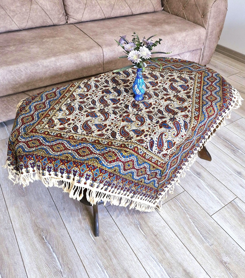May include: A white tablecloth with a paisley pattern in shades of blue, red, and gold. The tablecloth has a fringe border and is draped over a coffee table.