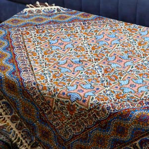 May include: A rectangular, intricately patterned textile with a cream-colored fringe. The design features a repeating pattern of blue birds, orange flowers, and geometric shapes on a pink and cream background. The border is a mix of blue, orange, and gold.