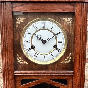 May include: A dark wood mantel clock with a white face and gold Roman numeral hour markers. The clock has a decorative design on the top and a glass window to view the clock face.