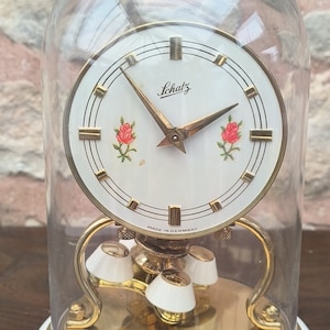 May include: A vintage gold-toned Schatz pendulum clock with a white face and black Roman numeral hour markers. The clock has a glass dome and a base with a white and floral design. The clock face features three red roses and the text "Schatz Made in Germany".