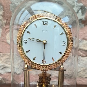 Può includere: Un orologio a pendolo Kundo dorato con un quadrante bianco e lancette nere. L'orologio è racchiuso in una cupola di vetro trasparente e poggia su una base dorata.