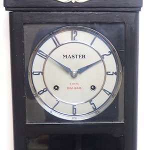 May include: A black wooden wall clock with a white face and silver hands. The clock face says "MASTER 8 DAYS BIM-BAM".
