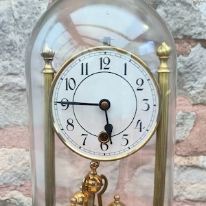 May include: A gold-toned pendulum clock with a white face and black hands. The clock is encased in a clear glass dome and sits on a gold-toned base.
