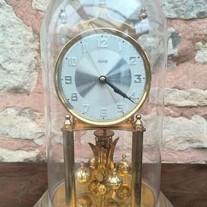 May include: A vintage Kundo clock with a silver face and gold-tone accents, encased in a clear glass dome. The clock features a pendulum and a round base. The clock is a decorative item.