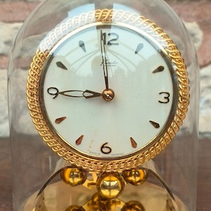 Può includere: Un orologio a pendolo dorato con un quadrante bianco e lancette nere. L'orologio è racchiuso in una cupola di vetro trasparente e poggia su una base dorata. Il quadrante dell'orologio indica "Rundo" e "Made in Germany".