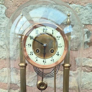 May include: A vintage brass clock with a copper face and black hands under a glass dome. The clock has a pendulum and is sitting on a wooden base.