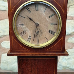 Puede incluir: Un reloj de madera antiguo con un cuerpo rectangular y una esfera redonda. La esfera tiene números romanos y un dial de segundos más pequeño. El reloj está hecho de madera oscura con incrustaciones decorativas y un bisel dorado.