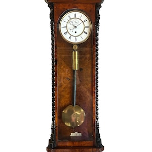 Peut inclure: Une horloge murale en bois avec un pendule. L'horloge a un cadran clair avec des chiffres romains et des aiguilles noirs. Le cadre de l'horloge est brun foncé avec des sculptures décoratives et un panneau de verre. Le pendule est de couleur or.