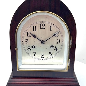 May include: A dark brown wooden mantel clock with a silver face and black numerals. The clock has a curved top and a rectangular base with gold-coloured feet. The clock face is framed with a gold-coloured metal.