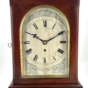 Puede incluir: Un reloj de chimenea de madera de caoba con una esfera de latón y marcadores de hora con números romanos. La esfera del reloj tiene un diseño grabado decorativo y el texto "The Grammarcy & Son 1. St. Michaels Alley Cornhill London 3716".