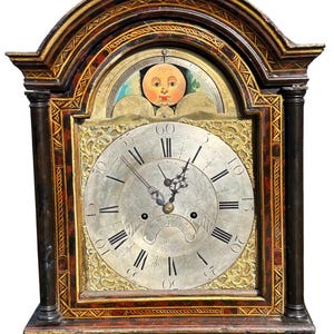 Puede incluir: Un reloj de pie antiguo con acabado estilo carey en color marrón oscuro y detalles pintados en oro. La esfera es plateada con números romanos negros, manecillas de metal ornamentadas y un indicador de fase lunar con un rostro.