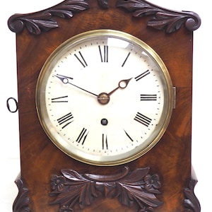 Könnte beinhalten: Eine Kaminuhr aus Holz mit einem geschnitzten Blumendesign an der Ober- und Unterseite. Das Zifferblatt der Uhr hat römische Ziffern und eine Glasabdeckung.